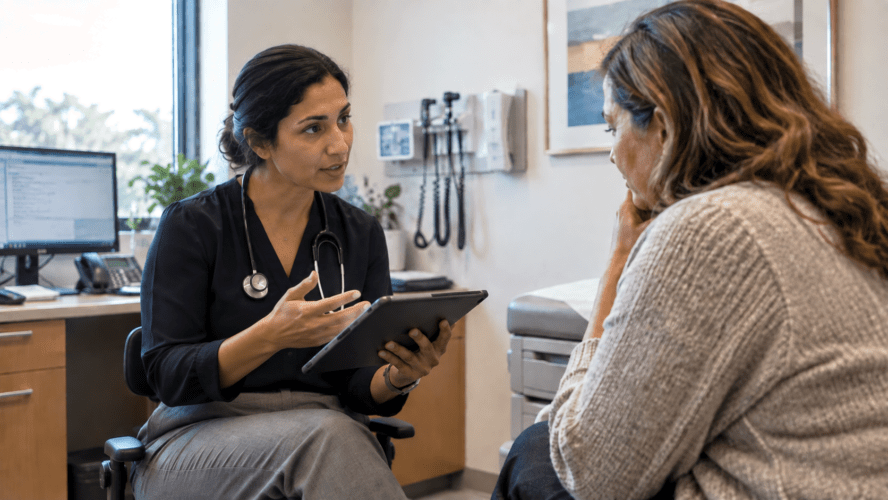 Doctor in business professional attire speaks with a patient during a clinic visit while holding a tablet, captured in a natural, candid moment.