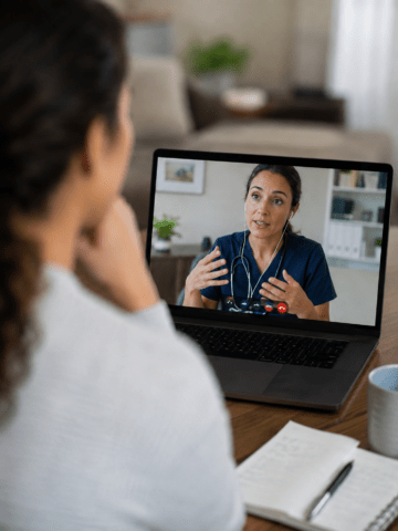 Patient participating in a telehealth appointment from home, viewed over the shoulder, with the doctor in navy scrubs in sharp focus on the laptop screen.