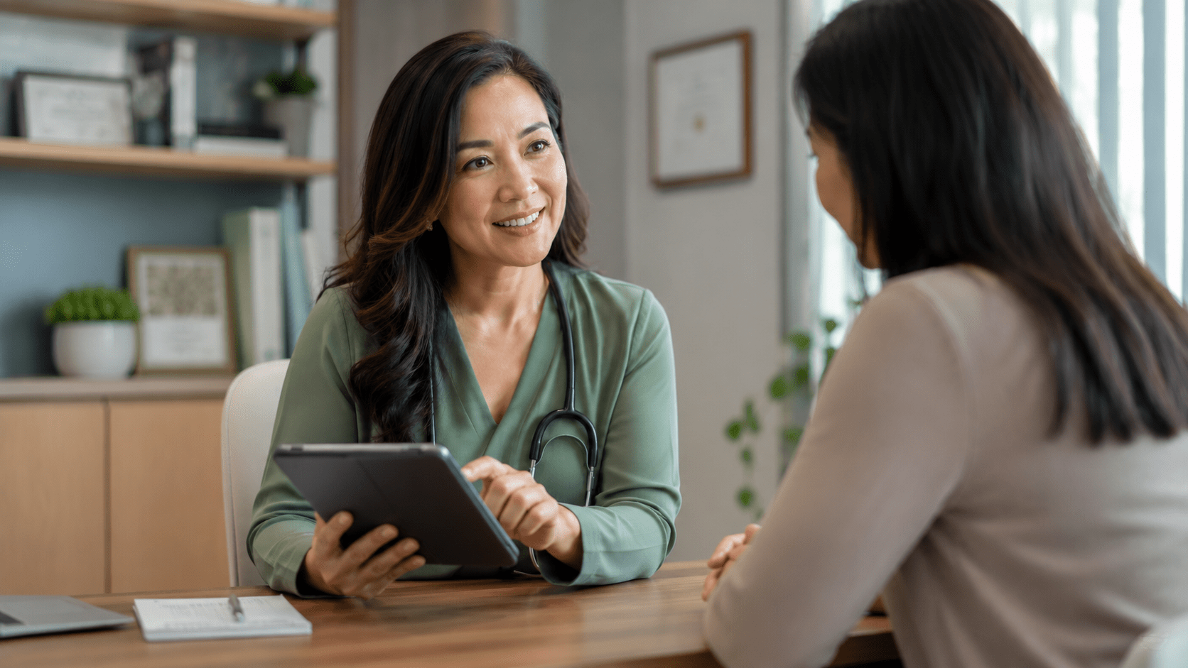 Female doctor wearing green using a tablet and talking to a female patient in a well-lit room