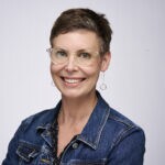 a woman with a denim jacket, short brown hair and classes smiling for the camera