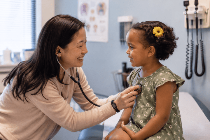 a brown haired female pediatric doctor standing with arms crossed in an exam room. the room has a picture of a giraffe and a turtle on the walls