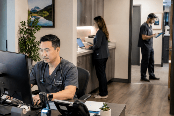 a busy doctor's office with doctors working at different stations using technology. all of the doctors are wearing dark scrubs