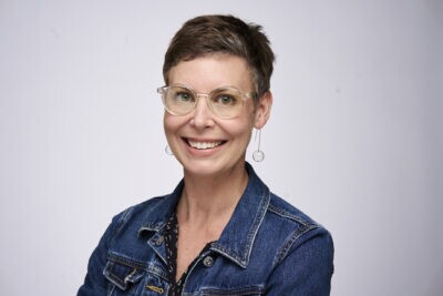 a woman with a denim jacket, short brown hair and classes smiling for the camera