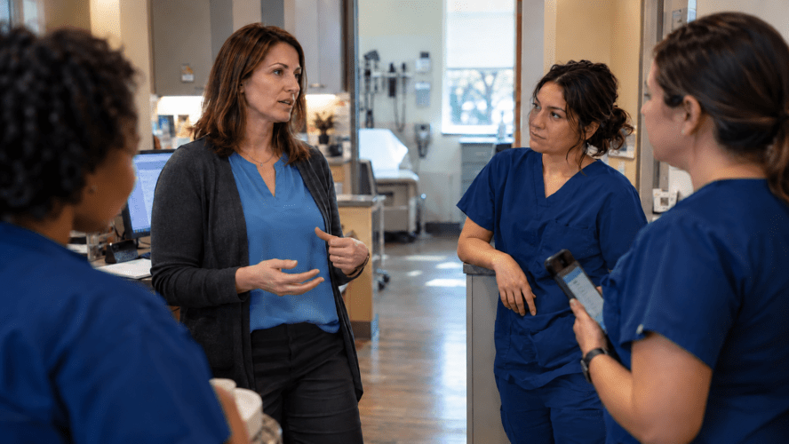 A female clinical leader talks with three staff members in matching navy scrubs in a doctor’s office, while one holds a tablet.